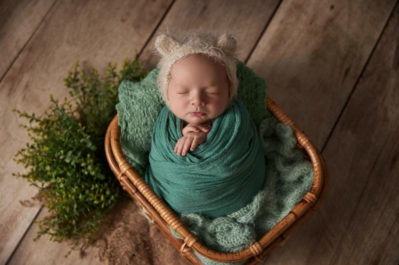Newborn miminko v košíčku s tyrkysovým wrapem a medvědí čepičkou.