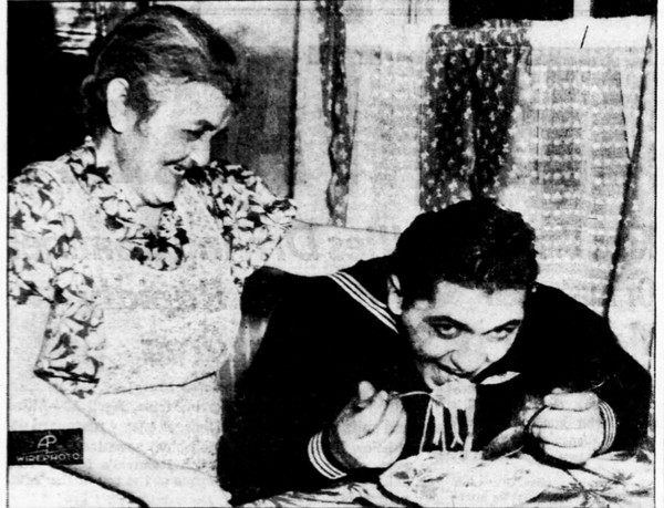 Basil Izzi sittin at a table with a fork-full of spaghetti in his mouth. His mother stands behind him with her hand on his shoulder.