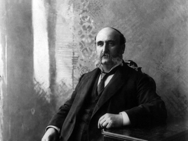 Ira Sankey sitting in a chair. He is wearing a black suit and has a bushy beard.
