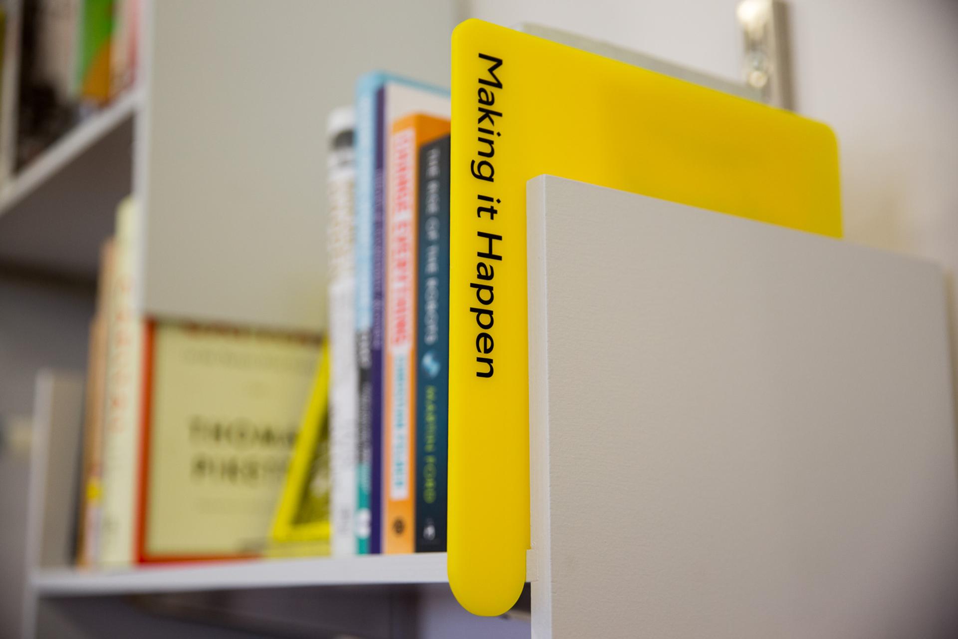 A grey bookshelf with a yellow divider with the words 'Making it Happen' and books in the background on the shelves, blurred out