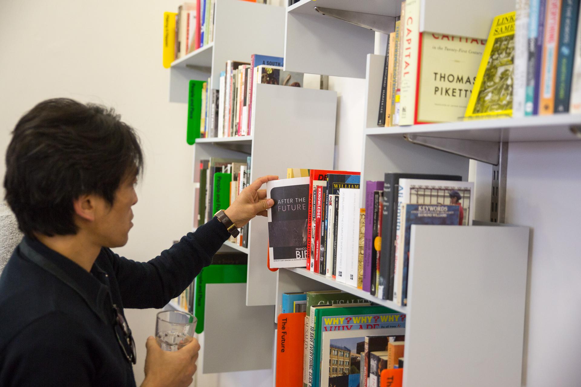 On the right, grey wallmounted shelves full of books. On the left, a man with dark hair and shirt, holding a glass and browsing the books
