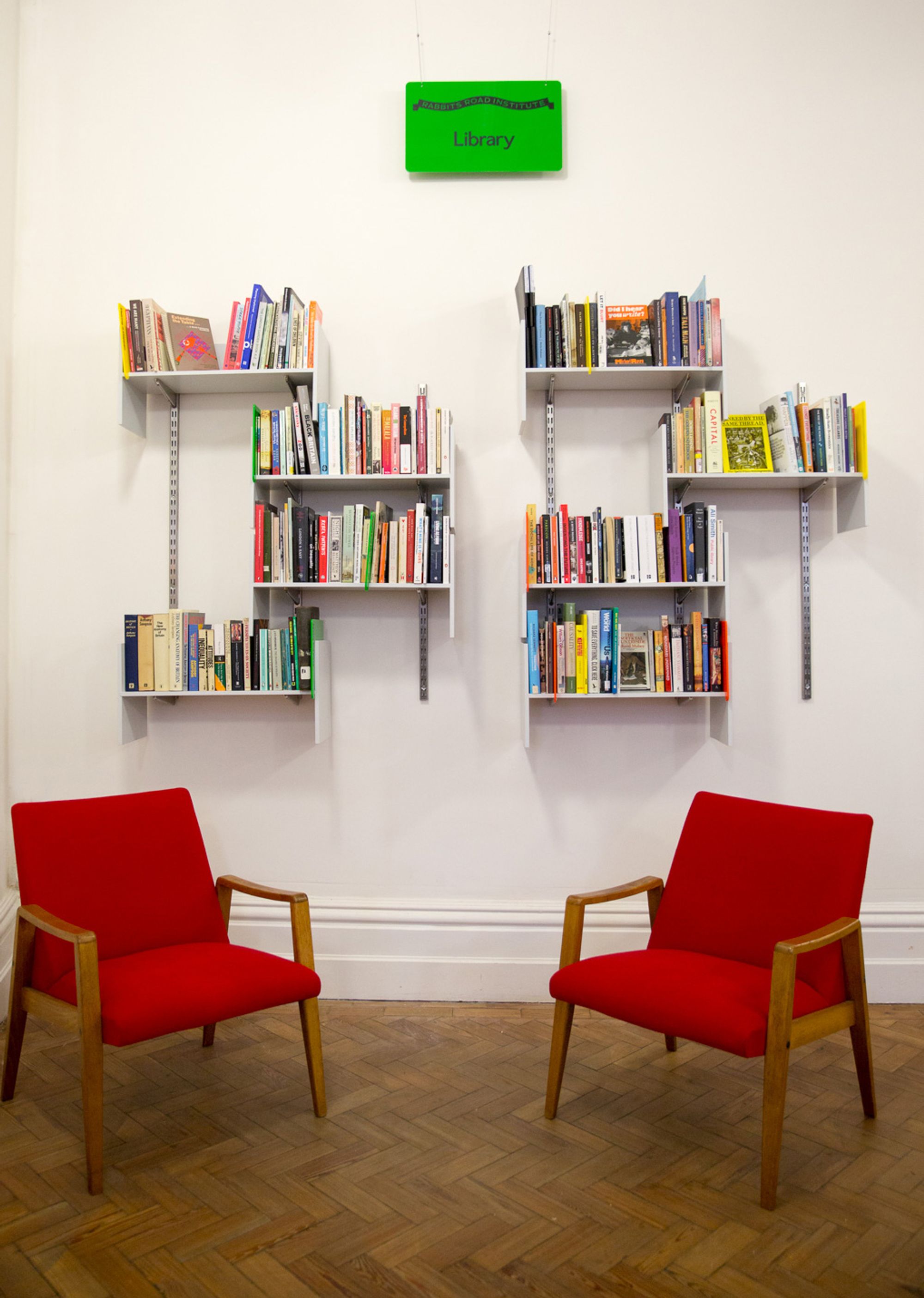 Eight wall mounted shelves full of books on a white wall; a green sign with the word 'Library' hangs above; in the foreground, two red midcentury lounge chairs.