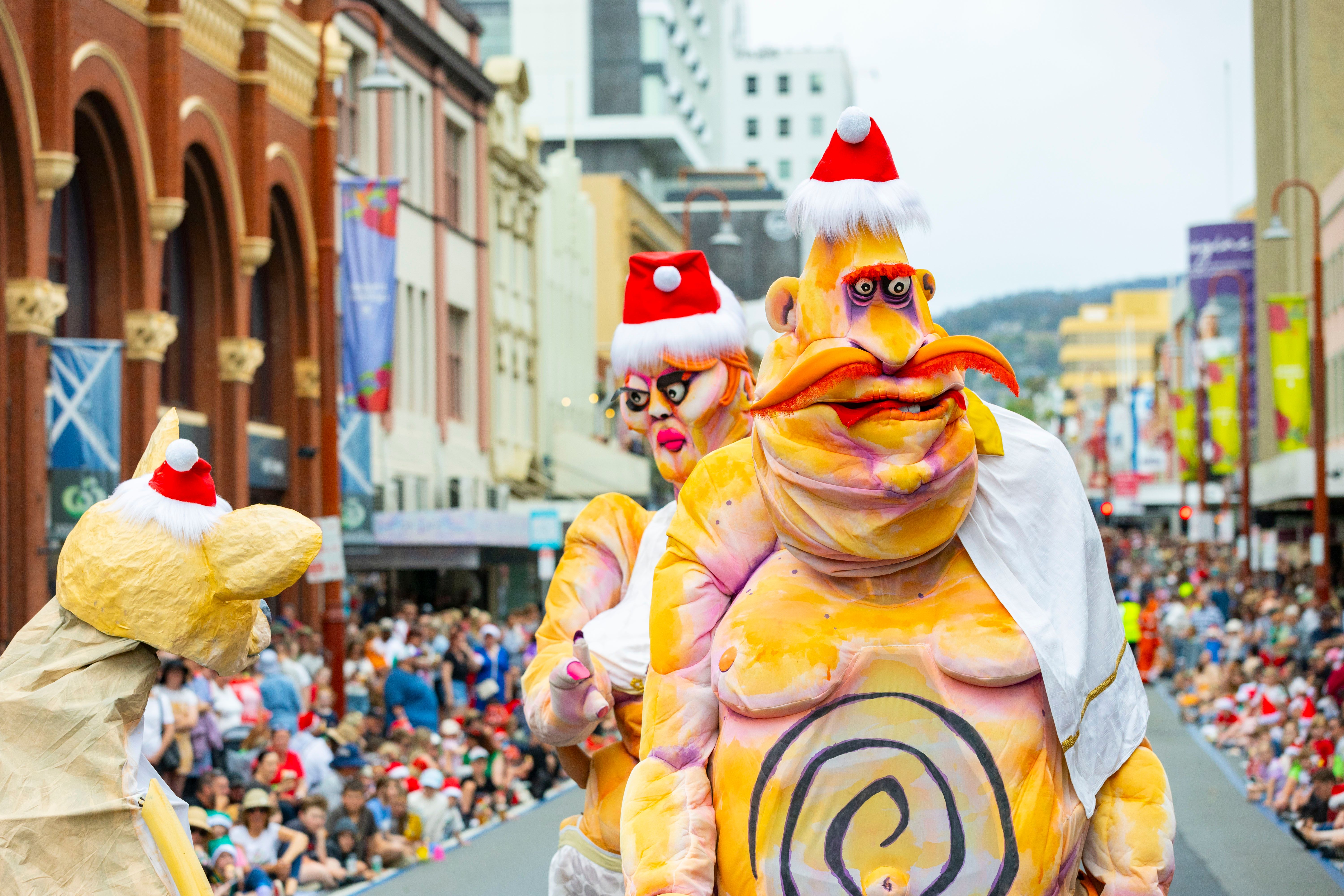 Two giant roving puppets, named Ma and Pa Ubu who both have exaggerated features, Christmas hats on their heads, one has a large orange moustache, the other bright neon pink lips, are walking in the City of Hobart Christmas Pageant, 2024.