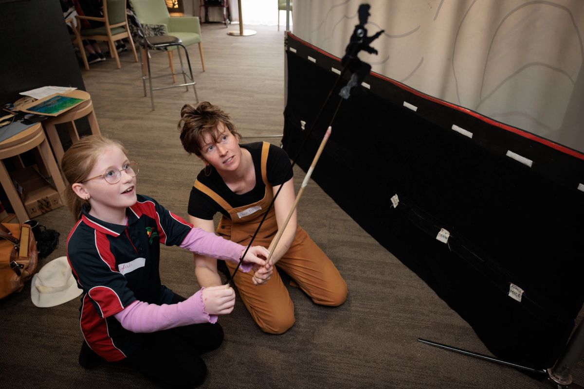 Two people, one a young person the other an adult are showing shadow puppets in front of a screen.