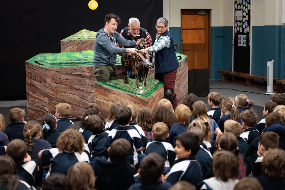 A puppetry performance is being performed in front of a group of school children. This performance looks like a green mountainous landscape, three performers, a skeletal wallaby like creature, a black background and a yellow moon in the distance.