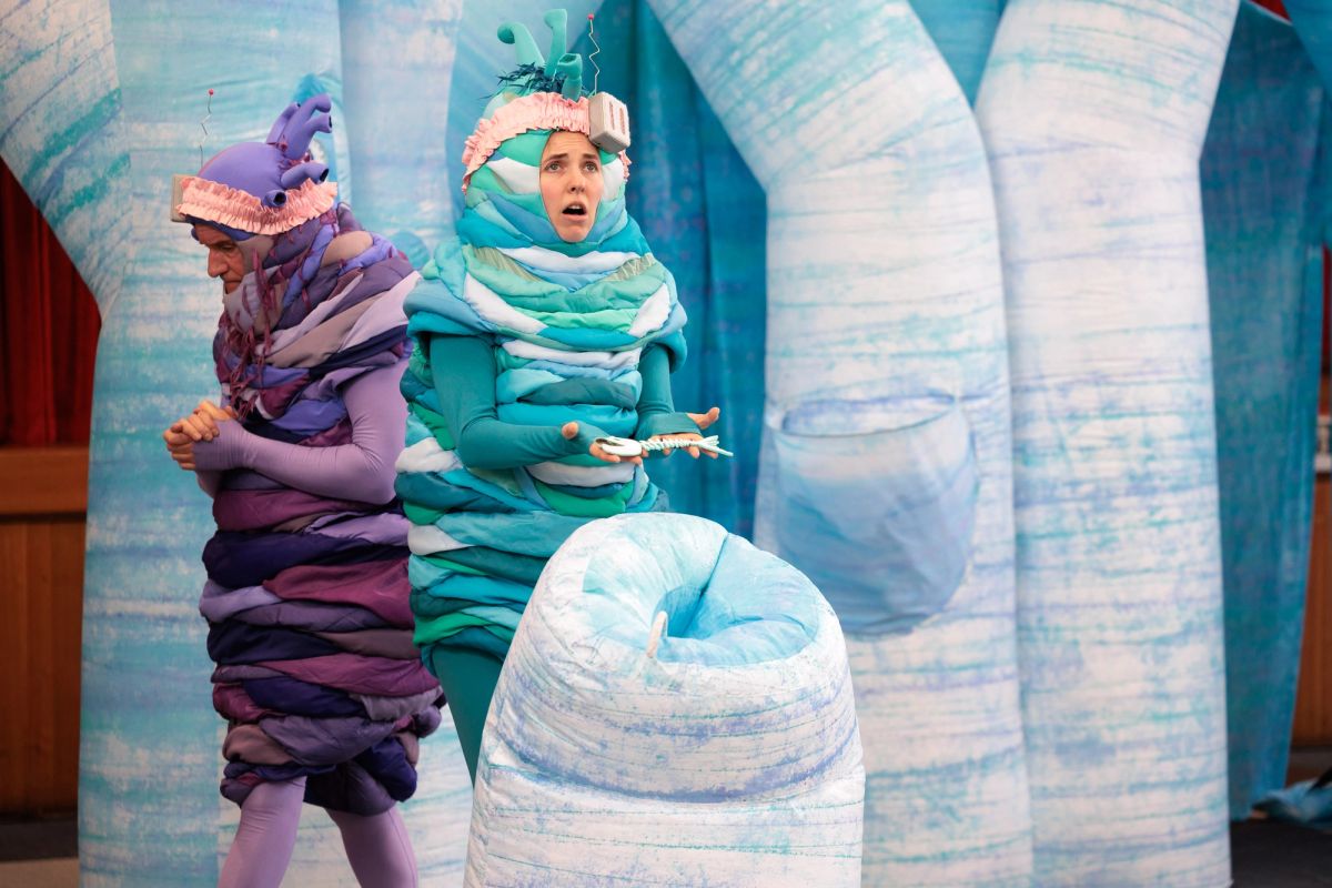 In Space Neighbours on Uranus, the teal alien holds a small white fish skeleton over an icy well, ready for a solemn farewell. Beside her, the purple alien bows his head, hands clasped. Both wear frilly pink headbands.