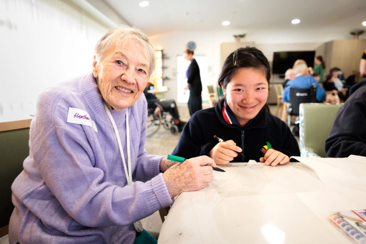 Two people sit at a table, pens/textas in hand one is an elderly person, the other a young person, they seem to be in a common room