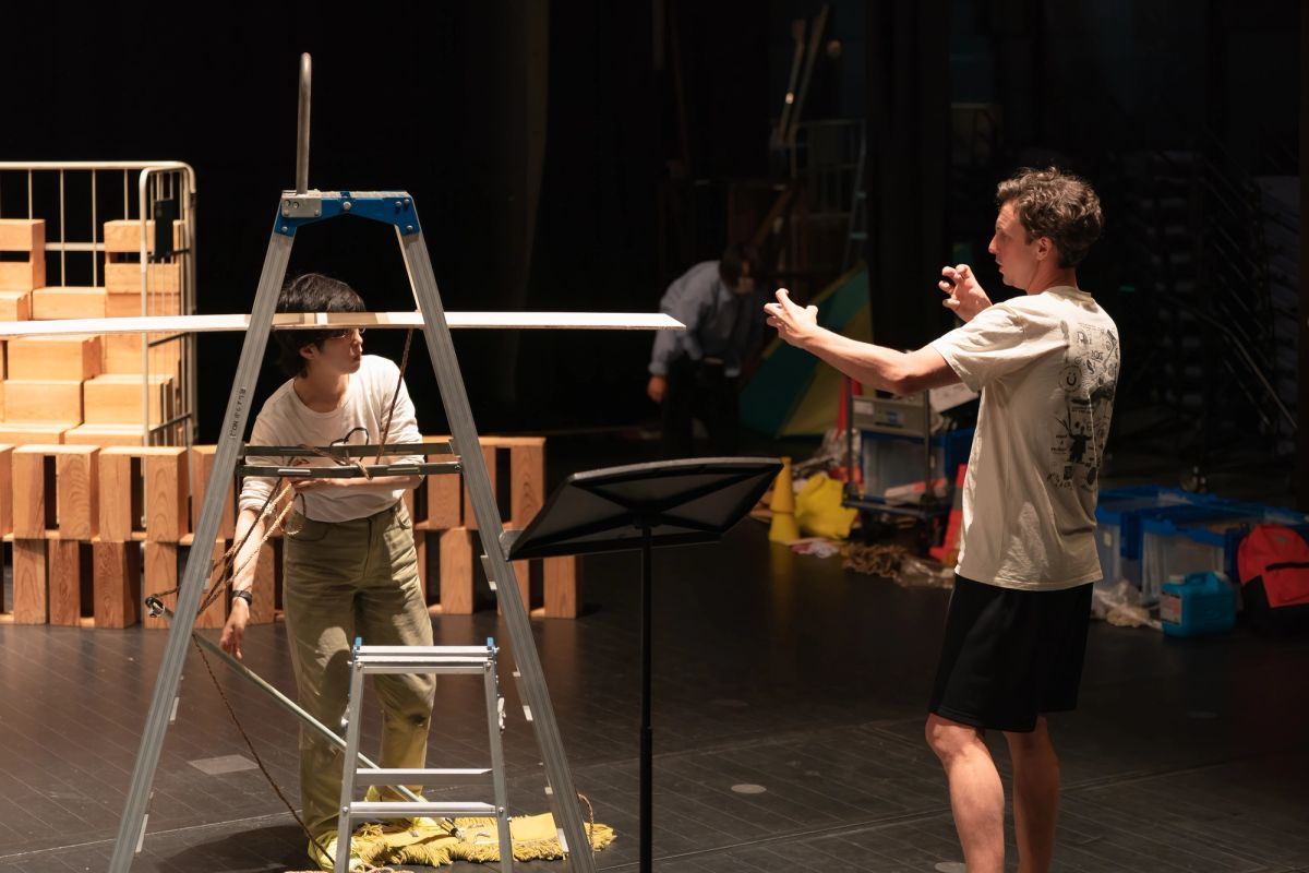 A person stands in a blackbox theatre, there are numerous items near them, including another person behind a ladder.