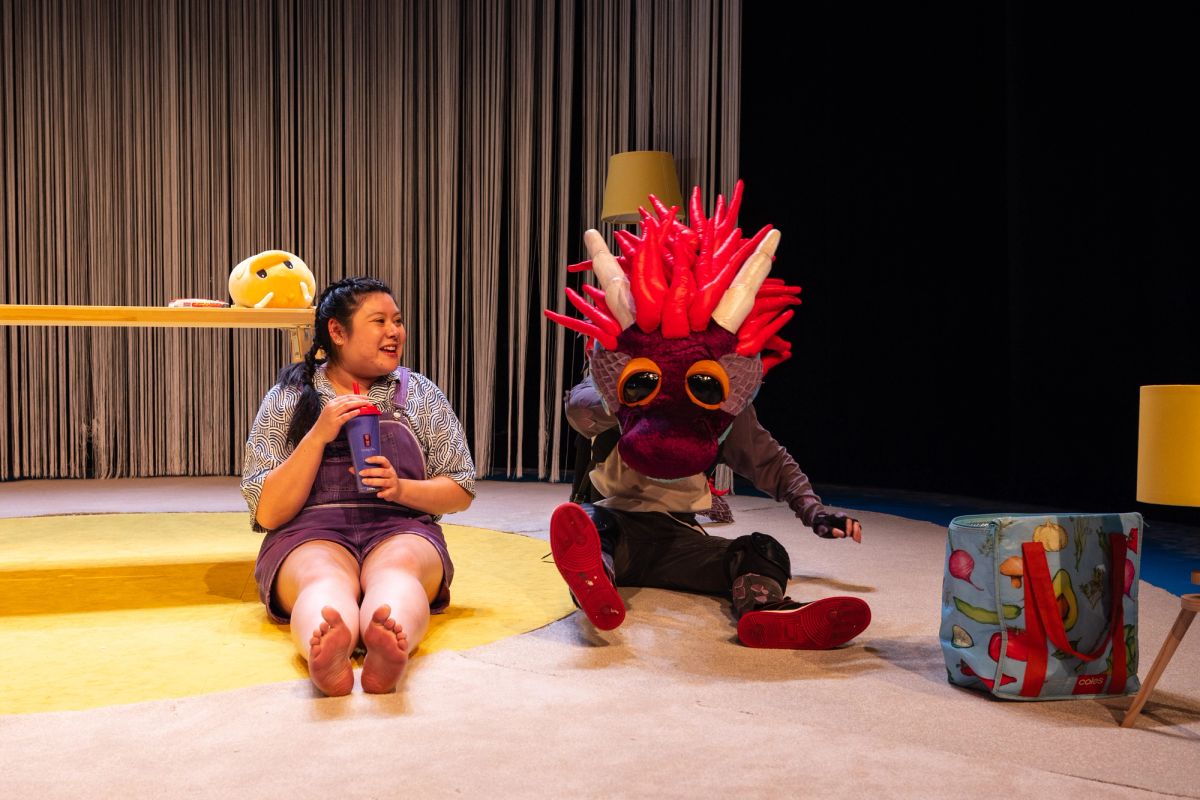 Chi sits on the floor smiling, holding a drink, beside dragon puppet Mizu, whose head is large and bright with red horns. The moment, from The Story of Chi performed at Adelaide Festival Centre as part of OzAsia Festival 2024