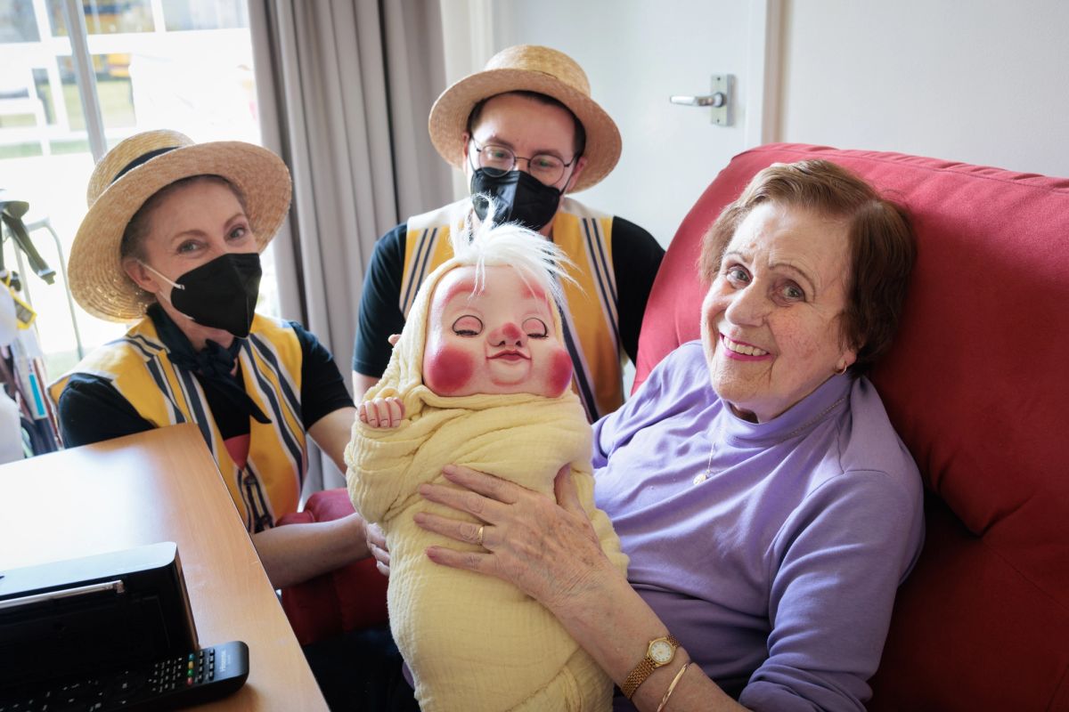 three people are in shot, all looking and smiling at the camera, one person is elderly and is holding a puppet baby, the other two are dressed in matching yellow and blue striped uniforms with boater hats and black face masks