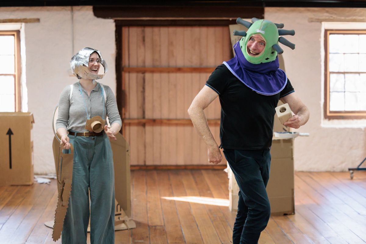 Two people rehearse in a room with cardboard set pieces. One person wears a cardboard helmet and holds a cardboard prop, while the other wears a large green headpiece with protruding shapes and carries a small cardboard item.