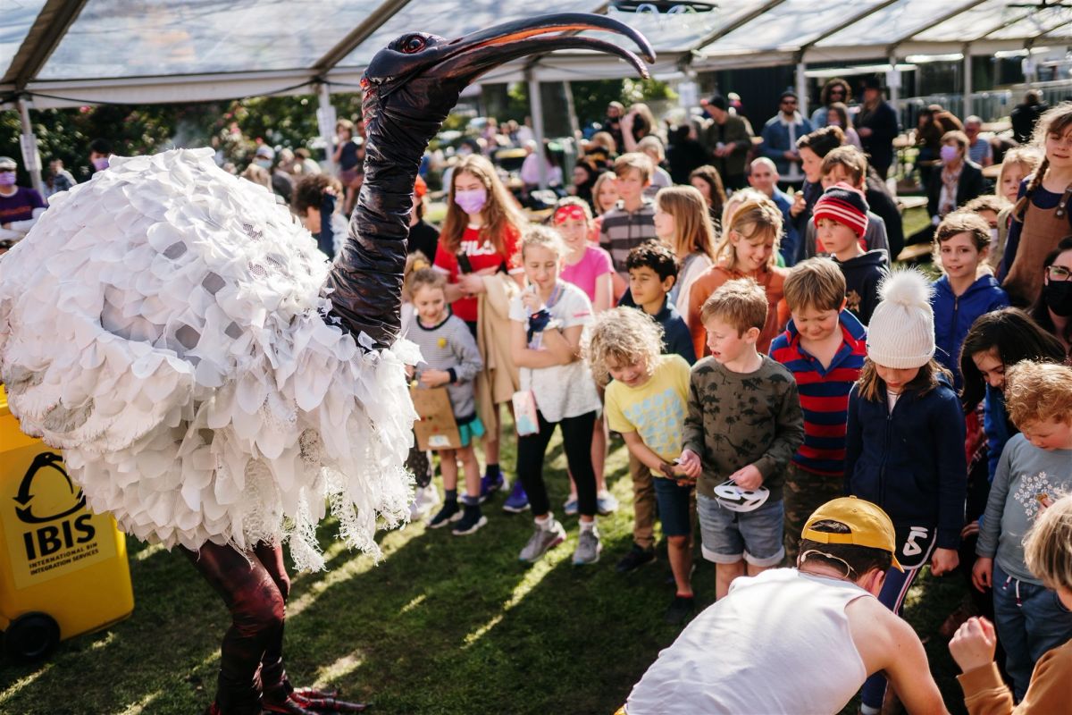 A larger than life size Ibis bird puppet, a white feathered body with long black legs and a long black neck and beak stands amongst a group of children at a festival.