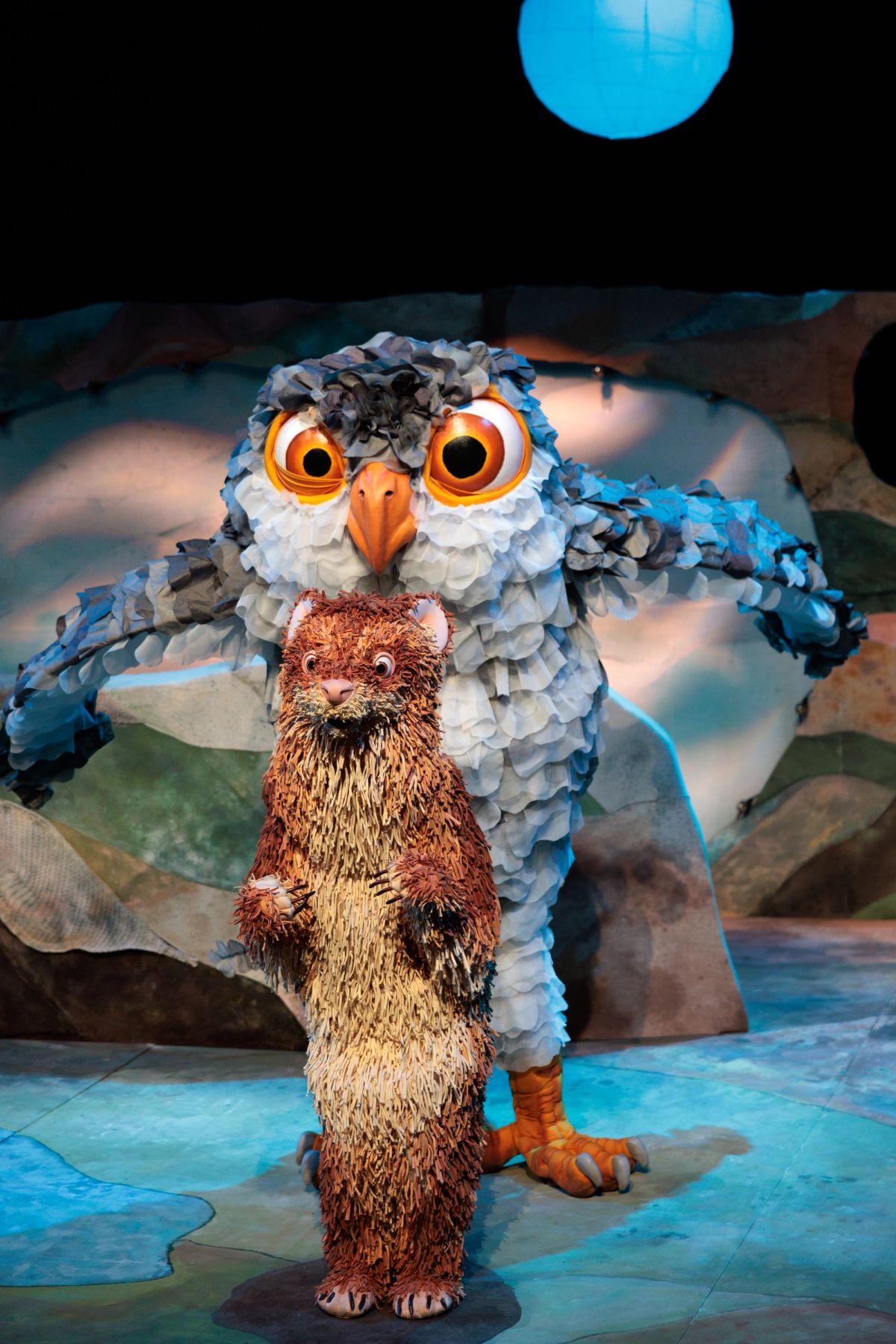 Feathers, a feathery-tailed ferret puppet, stands in front of a large owl puppet with wide orange eyes and outstretched wings, the mood is ominous and Feathers the ferret looks a bit scared. They are both on a painted stage set in Feathers at Theatre Royal Hobart.