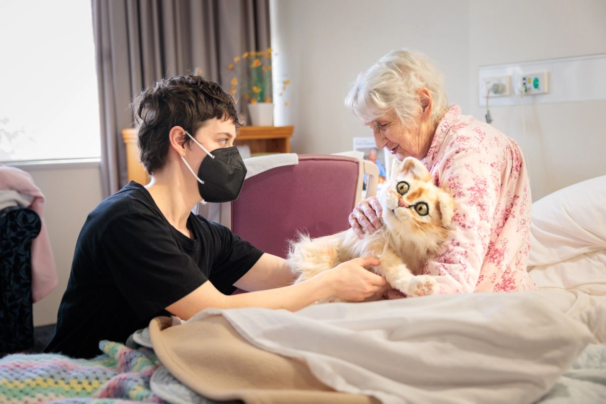 An elderly woman sitting on a bed holds and pats a cat puppet, there is another person puppeteering the cat puppet and