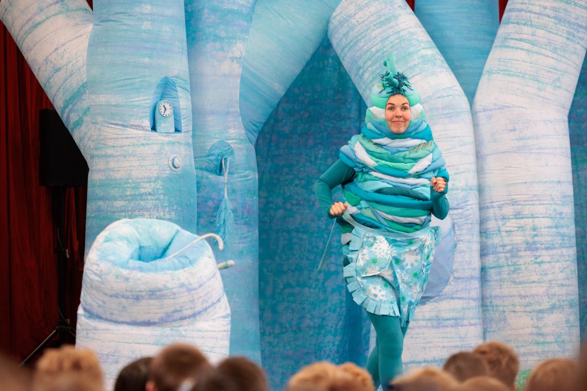 The teal alien in Space Neighbours on Uranus jogs across the stage with a cheeky smile, wearing a frilly snowflake apron. Tall icy blue set pieces rise behind her, with students' heads in the foreground.
