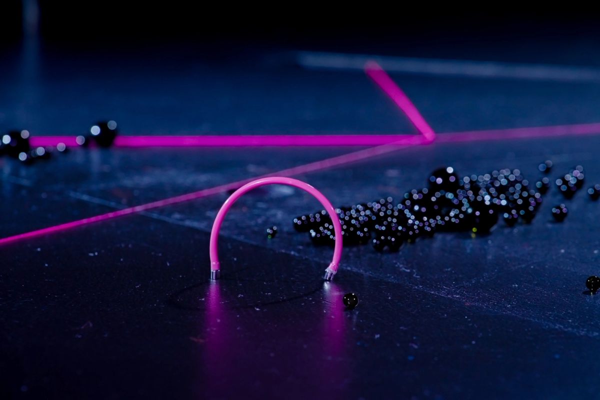 Dark stage with scattered oorbeez, blurred pink threads in the background, and a curved piece of pink electrical conduit in the foreground.