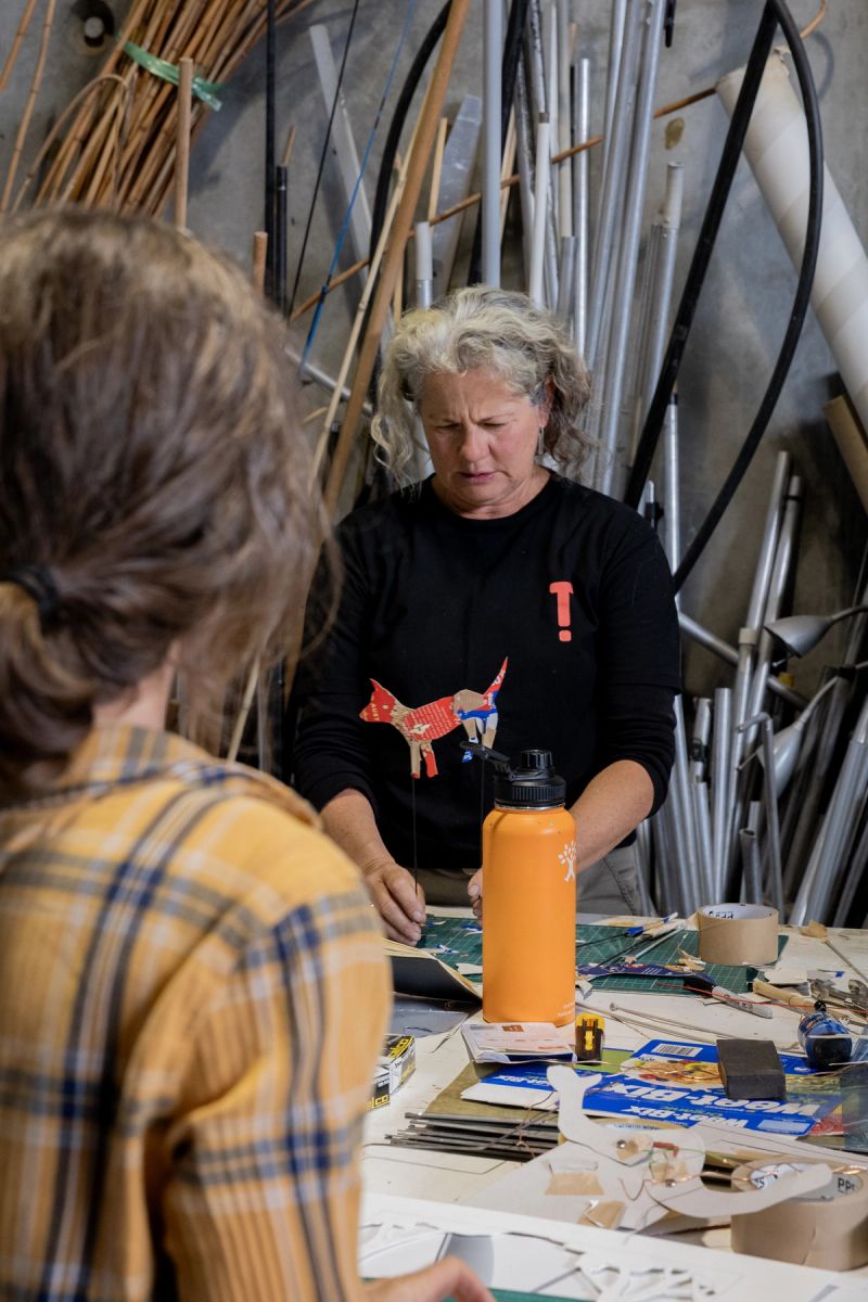 over the shoulder of one person we look ahead to another who is demonstrating how a dog shadow puppet works. These two people are in a workshop