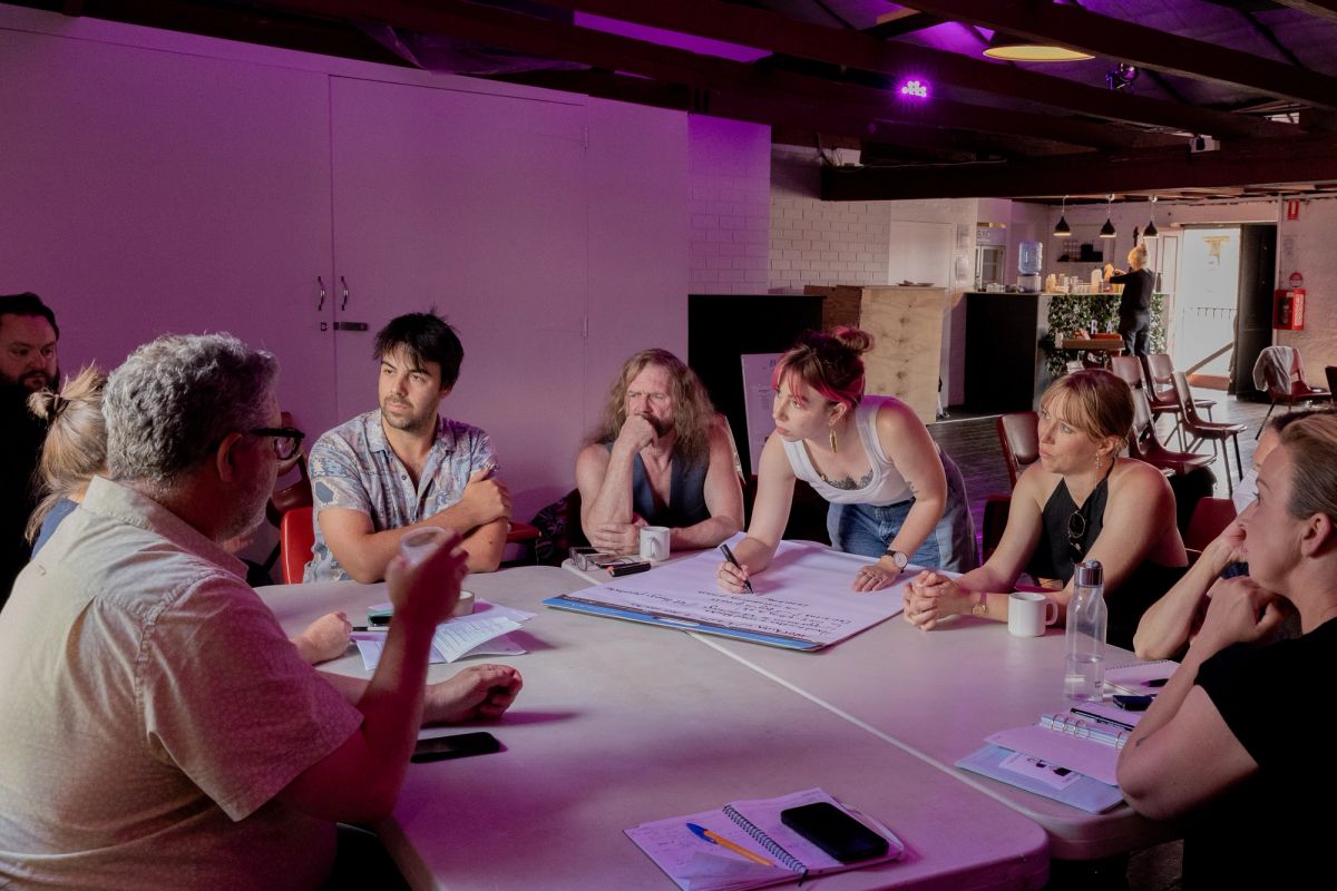 A group of 8 people are sitting and talking at a large table, they are all thinking and listening and one person with a large texta and large pad is writing down group thoughts.