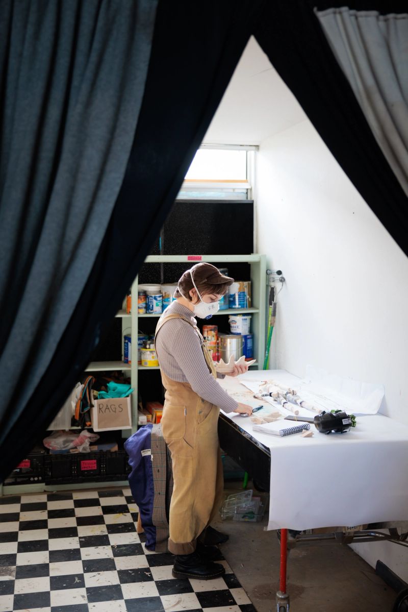 A person stands in a workshop, this workshop has black and white tiles, they stand over a table wearing a face mask.