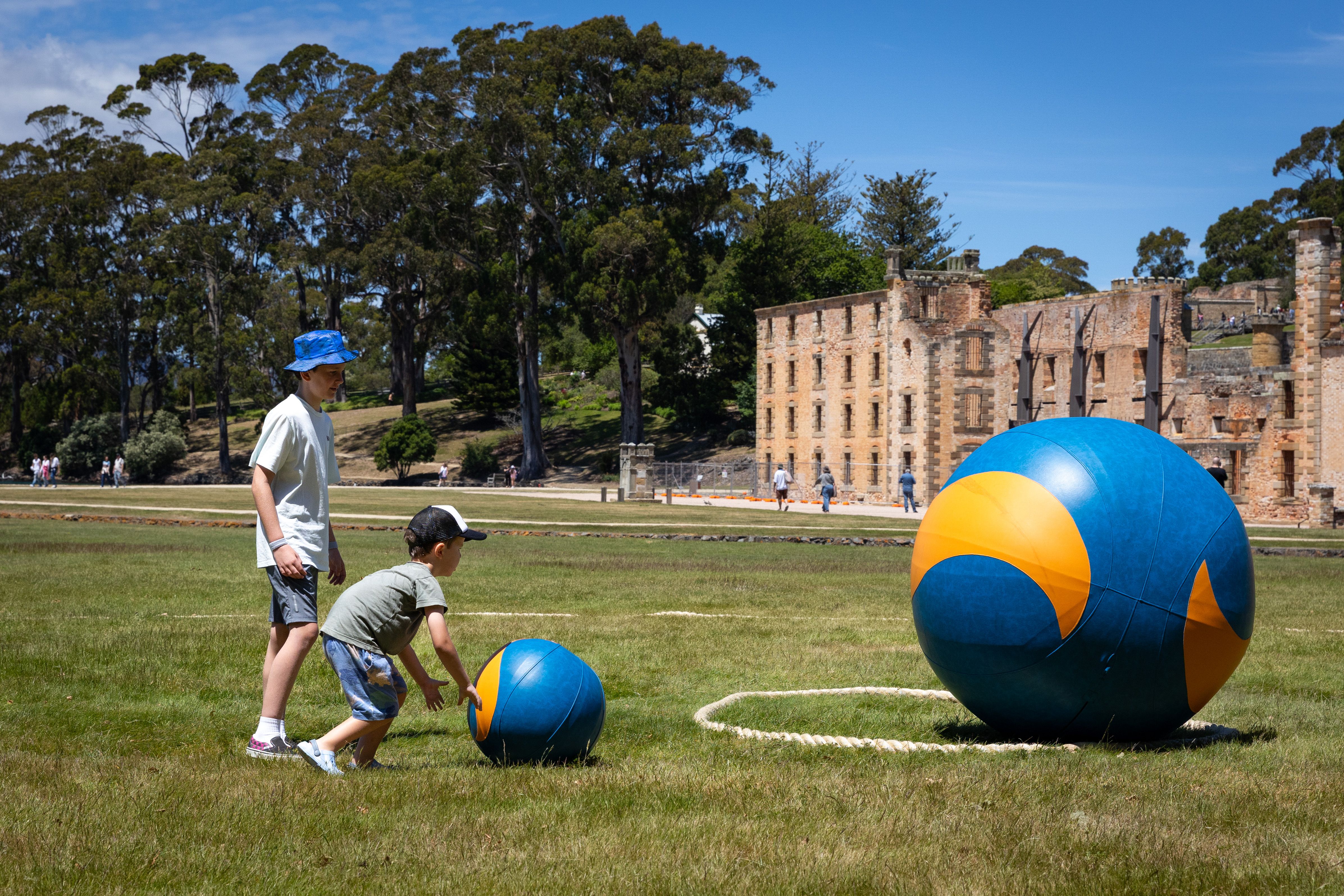 This is a bright sunny day, we see two young people playing a game of giant marbles, there are two marbles on lawn in front of the historic sandstone building of Port Arthur
