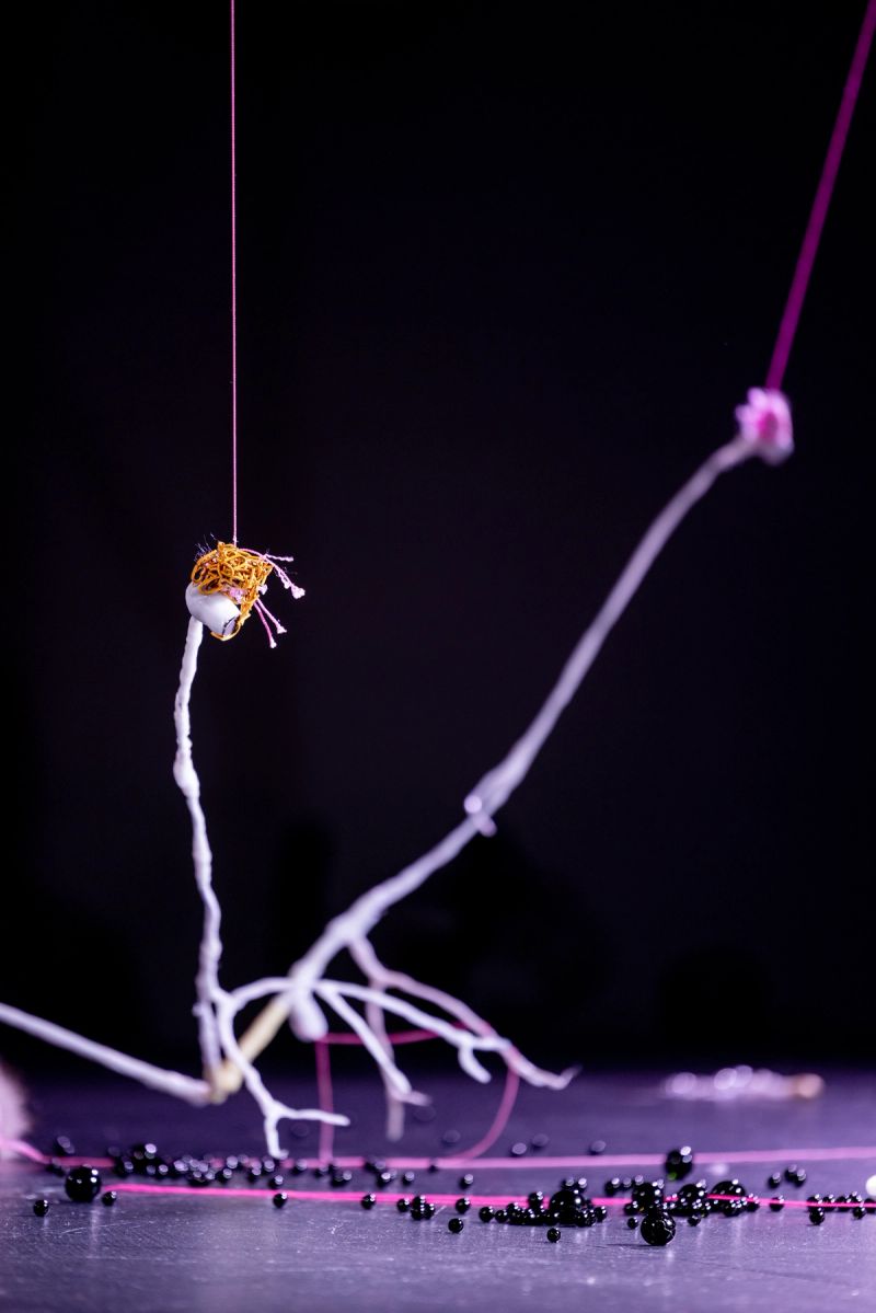 We see a low-lit stage with scattered oorbeez, in the foreground there is a white bone-like structure rising from the surface, and pink threads extending upward and orange/yellow bits on top of the bone.