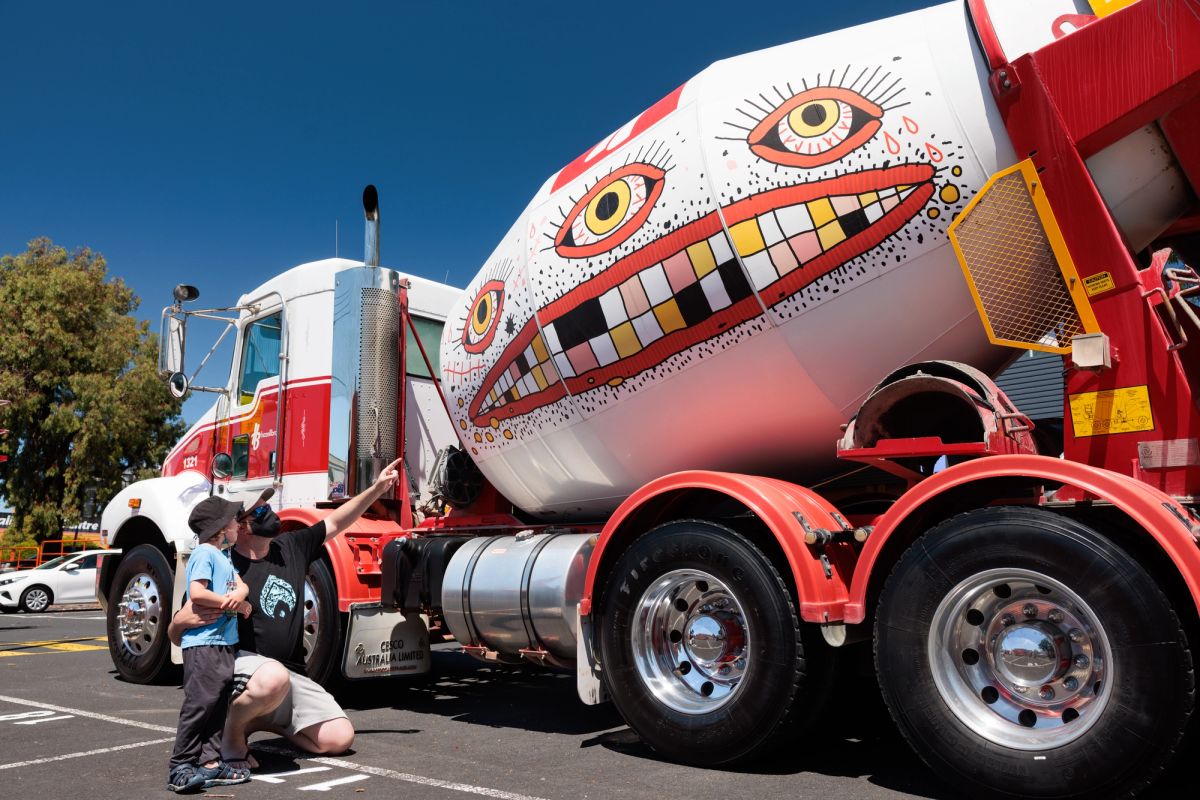 A large cement mixer is parked and an adult and young person look upon the artwork on the side of the truck, the artwork is a very large mouth, with teeth and three eyes