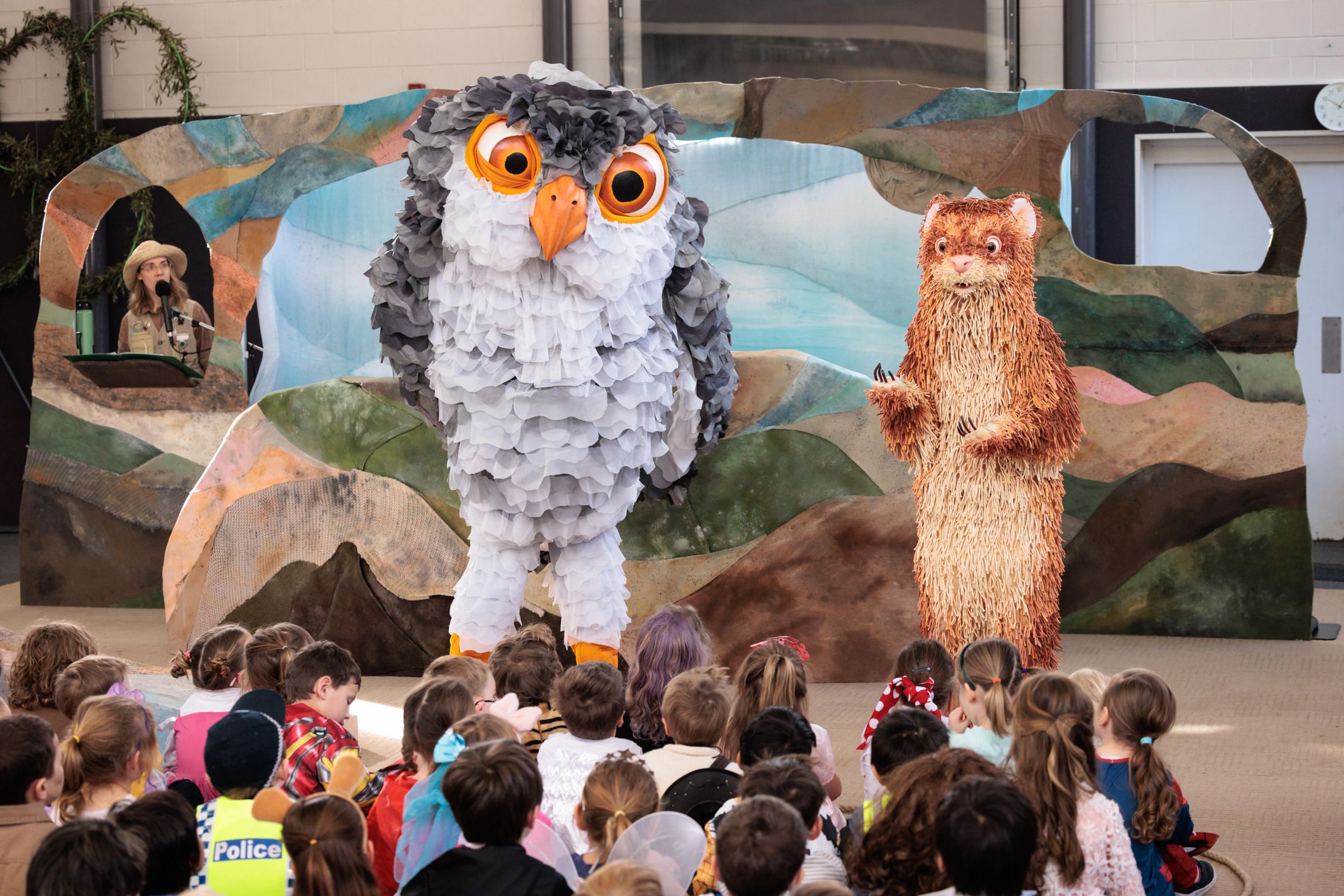 Feathers a giant ferret, a giant owl names Patsy are on stage in front of a group of school children, there is a set behind them made up of numerous muted colours.