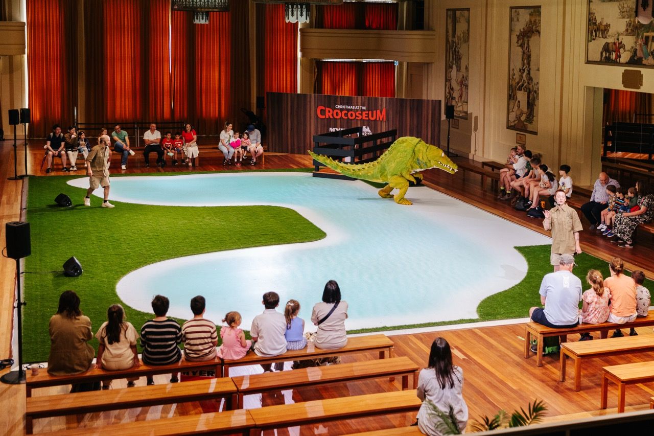 The Myer Melbourne Mural hall has been turned into a room with a performance in the centre, there is a crocodile show, with fake green grass, a blue watery like area and a puppet crocodile. around the edges of the room is seating filled with patrons watching the performance happen.