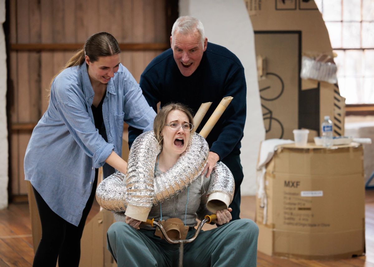 Two people stand behind a third person who is sitting on a small tricycle during a rehearsal. The seated person has flexible silver ducting and cardboard tubes wrapped around their shoulders and reacts with an open-mouthed expression, while the two behind them guide or steady them. Cardboard set pieces surround the group.
