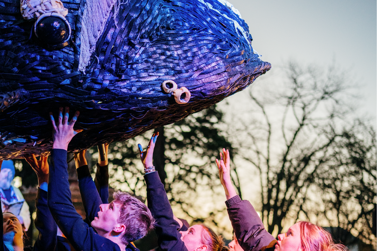 Hands raised, a group of young people lift and steady a large blue whale sculpture overhead at an outdoor festival as the sky fades into evening.
