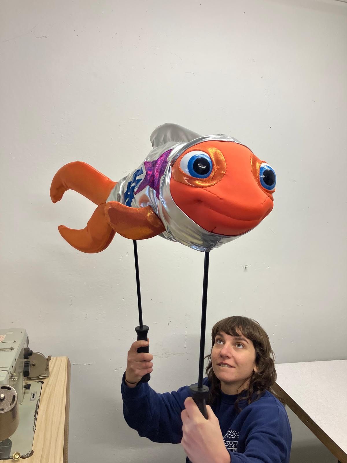 A person holds up via two long handles an orange fish puppet wearing a metalic space suit