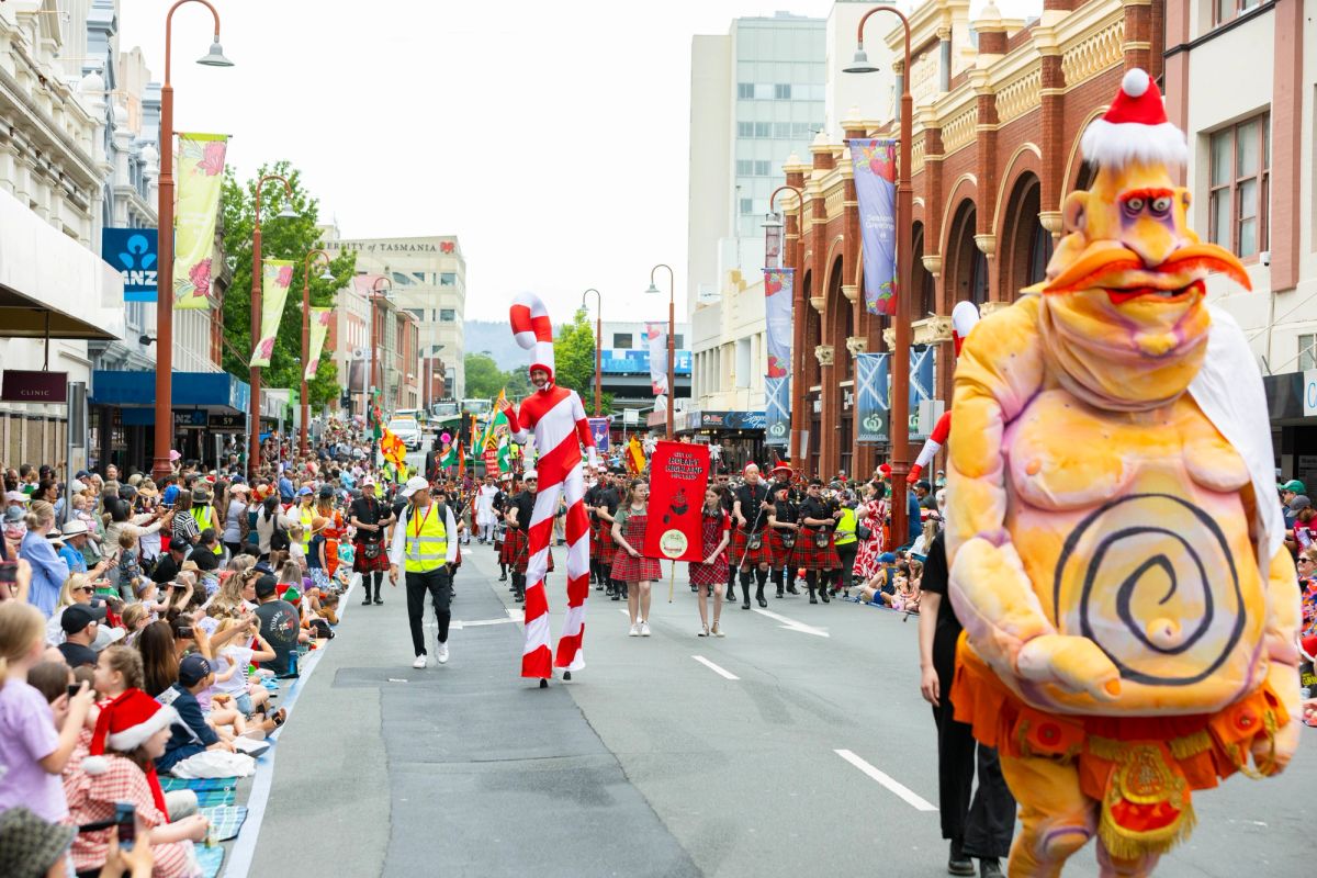Giant roving puppet, Pa Ubu who has exaggerated features like a Christmas hat on his head, and a large orange moustache is walking in the City of Hobart Christmas Pageant, 2024. In the background we see other pageant participants and the streets of Hobart lined with audience members.