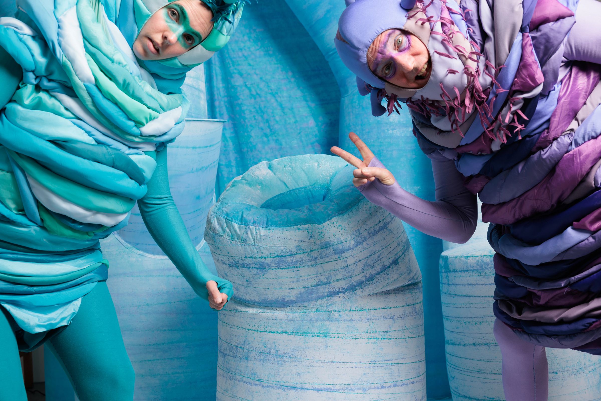 Two alien characters lean in toward the camera from either side of a large sculptural inflatable prop in pale blue. The teal alien on the left peers in with a cool, watchful expression, while the purple alien on the right flashes a peace sign and pulls a wide-eyed, comedic face. The entire scene is set within a textured blue environment.