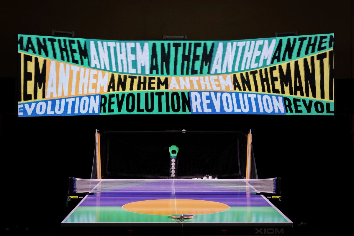 Set in front of a black background is a table tennis table. There is an larger than life tv screen directly in front of the player, it looks like three tv's together and in a mix of bright australiana colours the screen says ANTHEM ANTHEM REVOLUTION