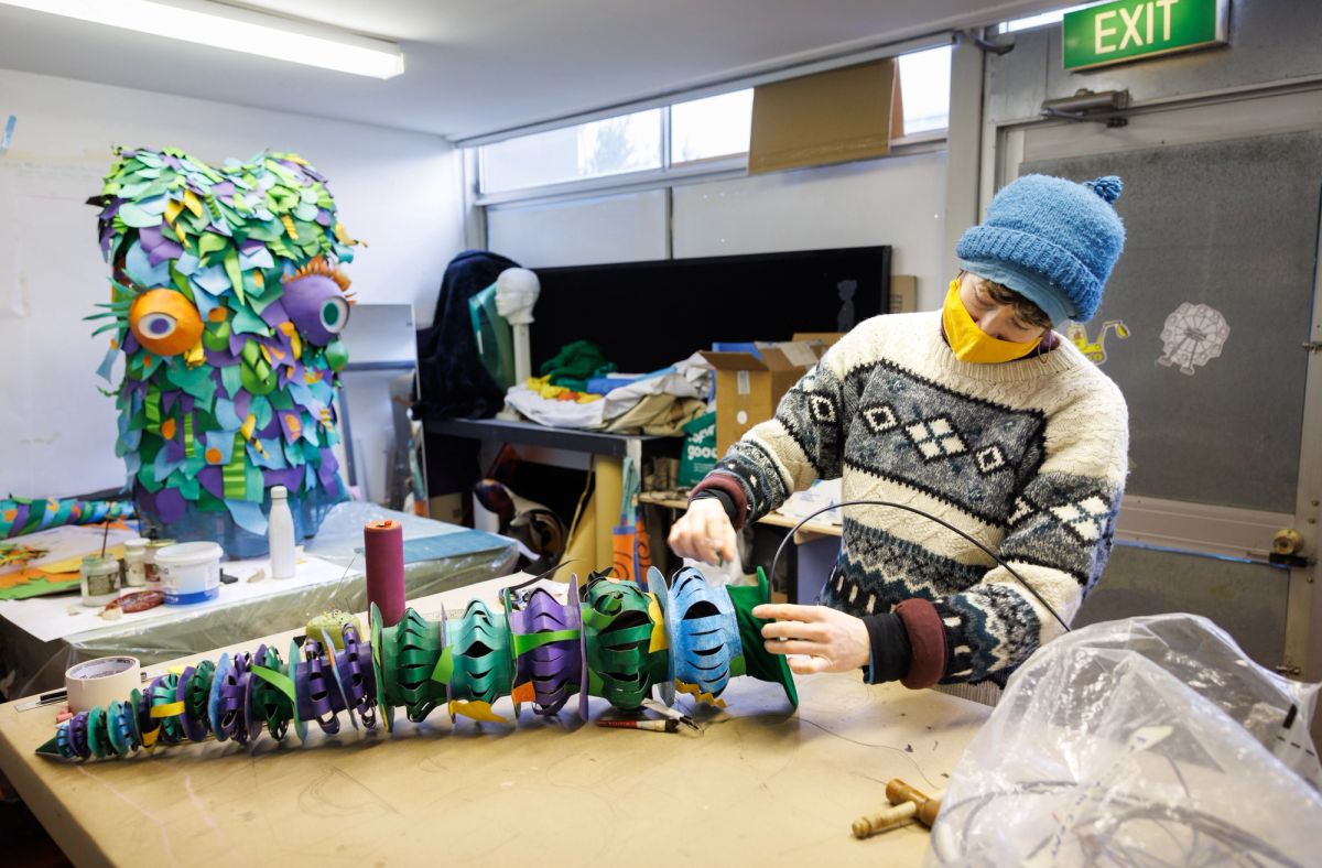 A person in a black and white woollen jumper stands at a work bench, they're working on what looks to be a colourful tentacle our of multicoloured paper. We know it's a tentacle because we see the body of the colourful octopus in the background