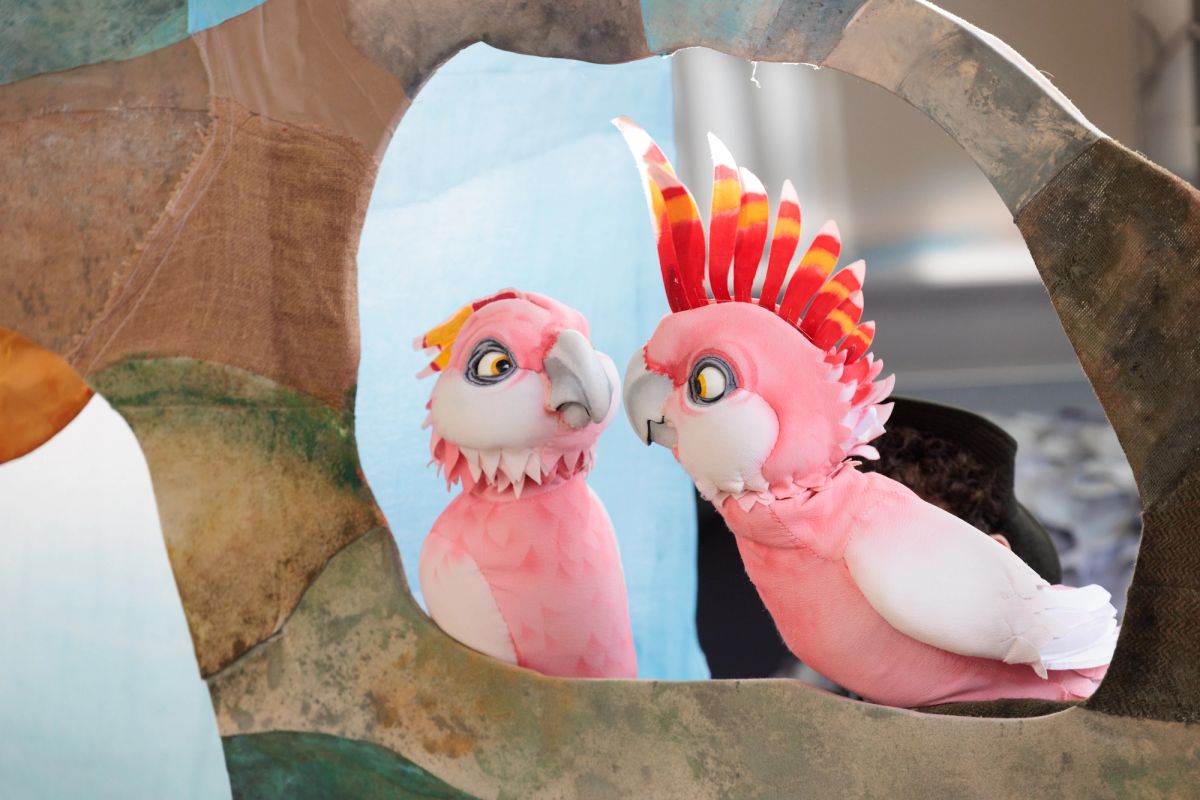 Two cockatoo puppets are looking at one another, one with it's crest up, they're pink, white and yellow in colour