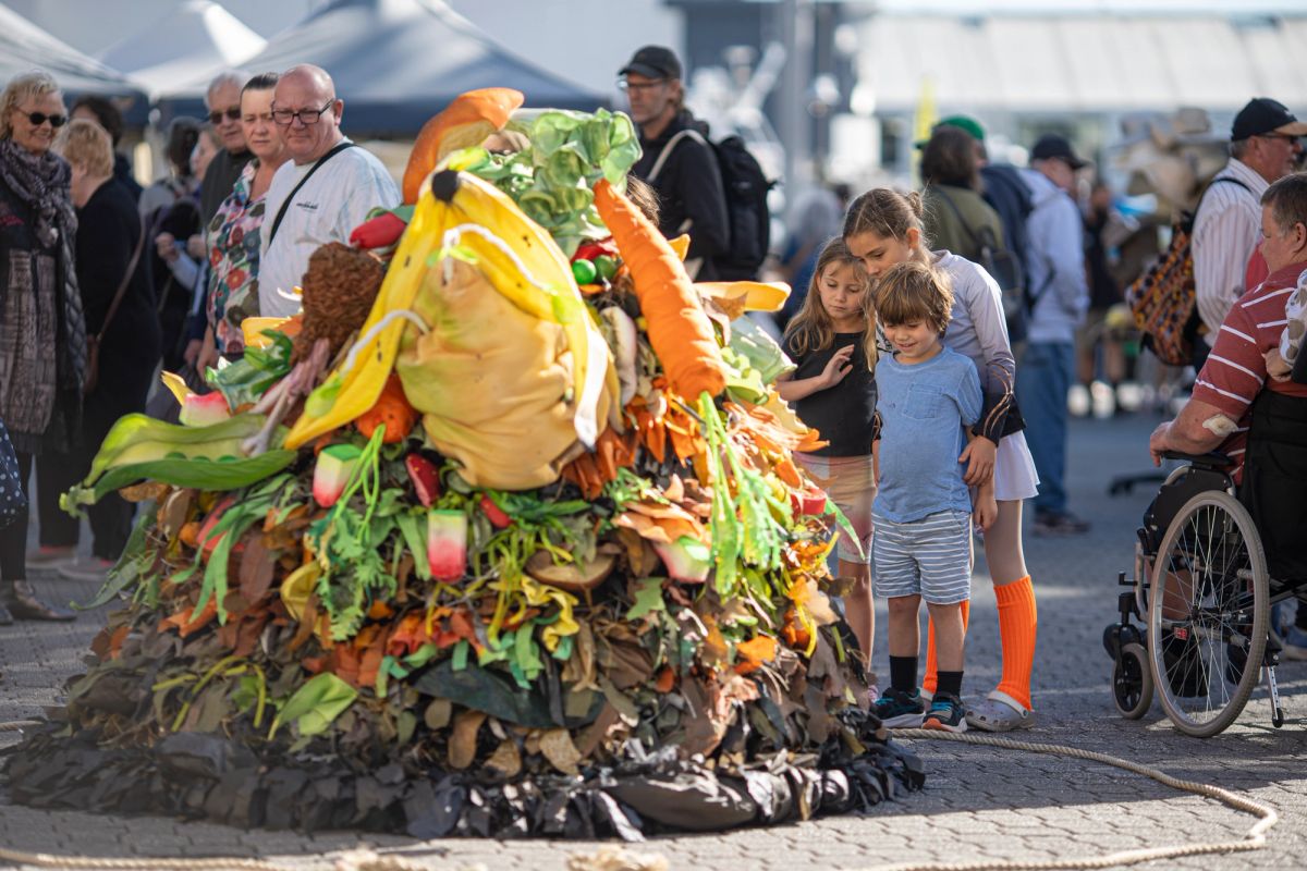 A pile of compost lays on the ground, but this isn't just any compost, it's a performance about to start. Children look on excited. In the compost heap is a visible carrot, watermelon, banana peel and a pod of peas.