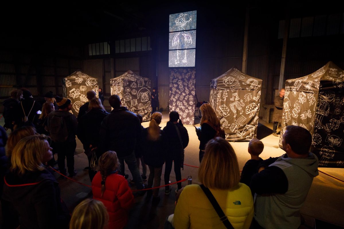 A group of people are standing and looking at four black marquees covered in white drawings as well as up high there are three tv screens showing a black background and white drawing