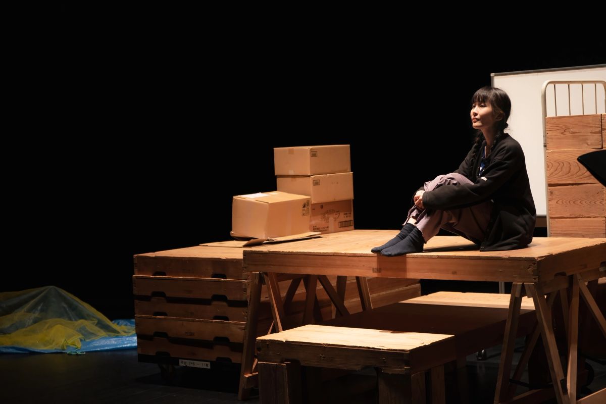 A person sits on top of a brown wooden table surrounded by brown boxes, this person is under stage lighting so everything else is in blackness