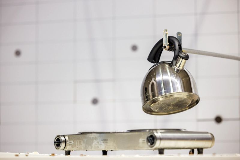 We see a variety of breakfast paraphernalia on a table, here we see a kettle and a hotplate operated by a long pole from a wall off in the distance.