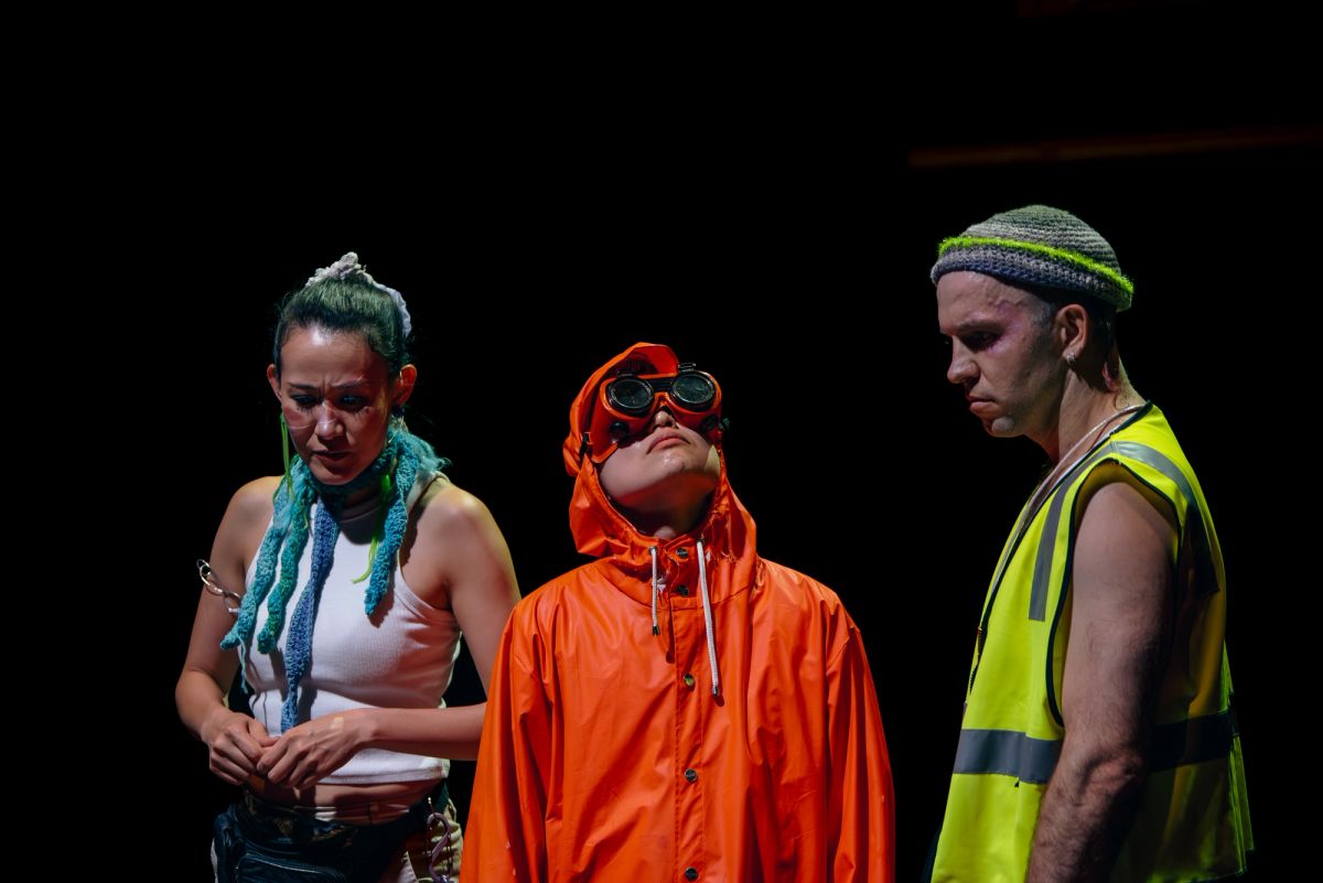 Three performers stand closely together in dramatic lighting. One wears a white tank top with blue scarves, looking down. Another wears an orange raincoat with large goggles, gazing upward. The third wears a high-visibility vest and a knitted cap, staring intently.