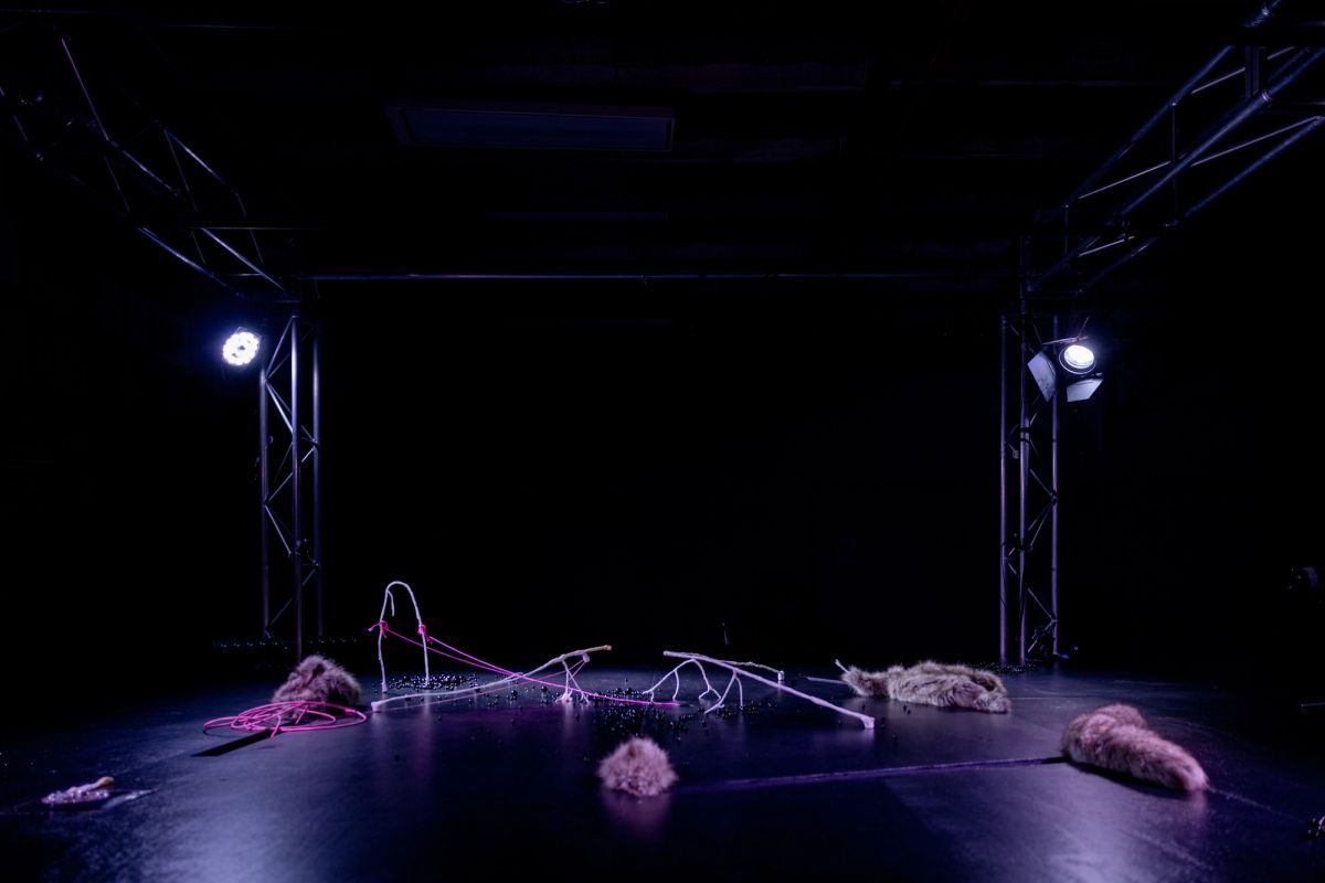 A wide angle of the Matter Era stage, we see on either side of the stage truss with big lights shining down, it's very low-lit, with oorbeez scattered across the surface, white bone-like structures, pink threads, and fur elements.