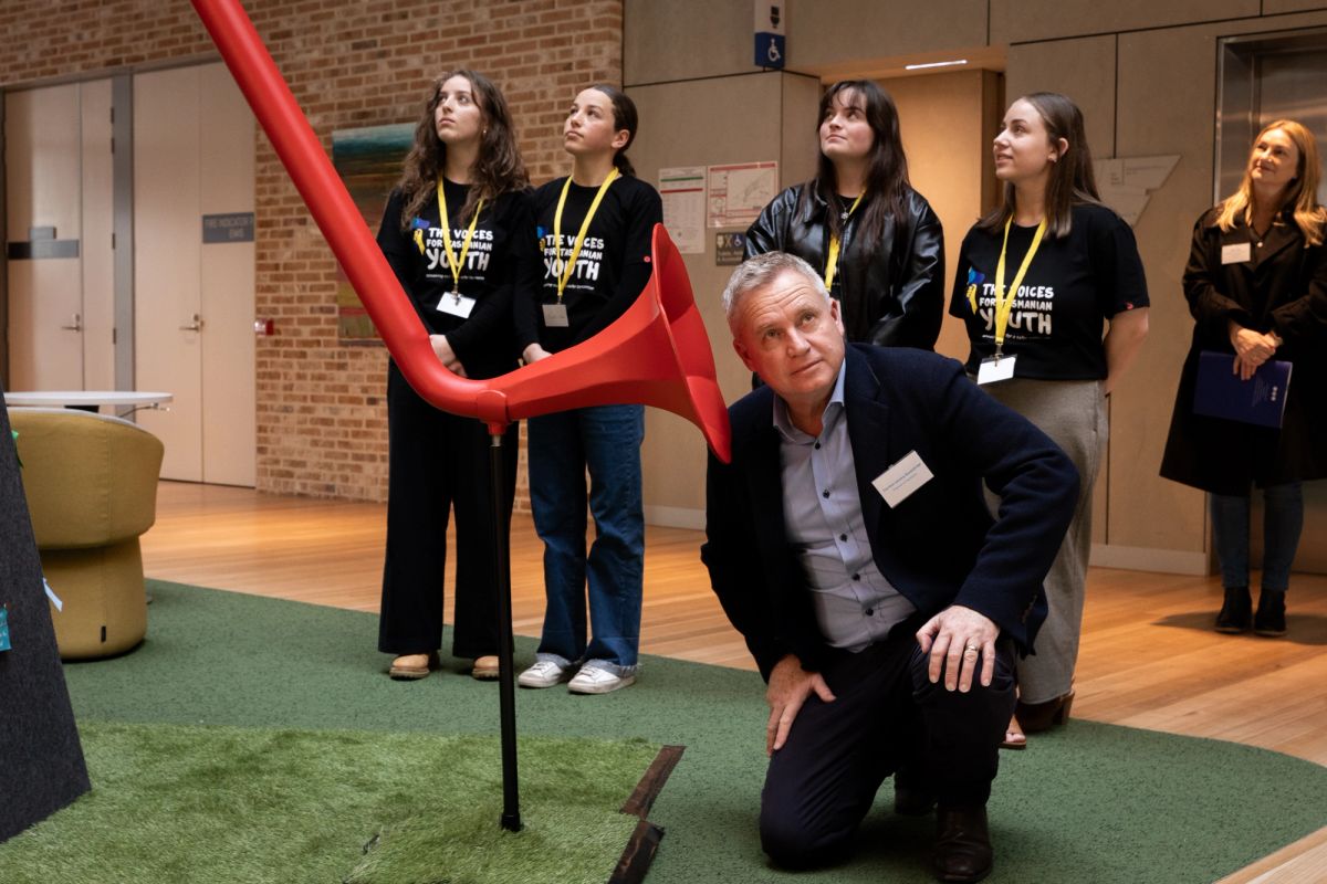 A person has their head to a red megaphone, at the other end of the megaphone is a young person, this person is on top of a small mountain which is used to amplify young peoples voices