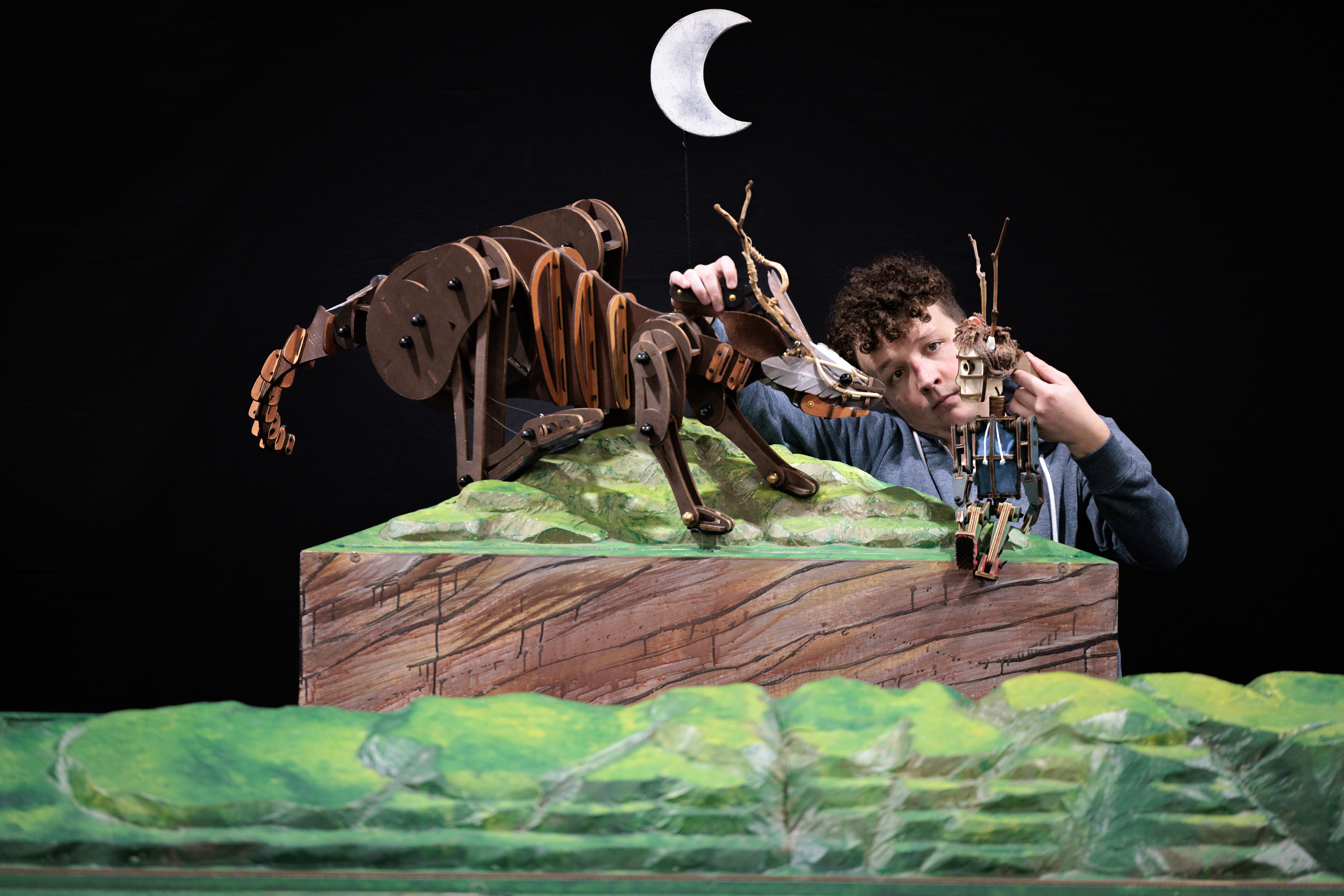 In front of a black background there is a person, holding two puppets, one puppet looks like a small person, the other like a skeletal wallaby. These puppets sit on top of a green mountain scape and there is a moon in the middle of the image in the black background.