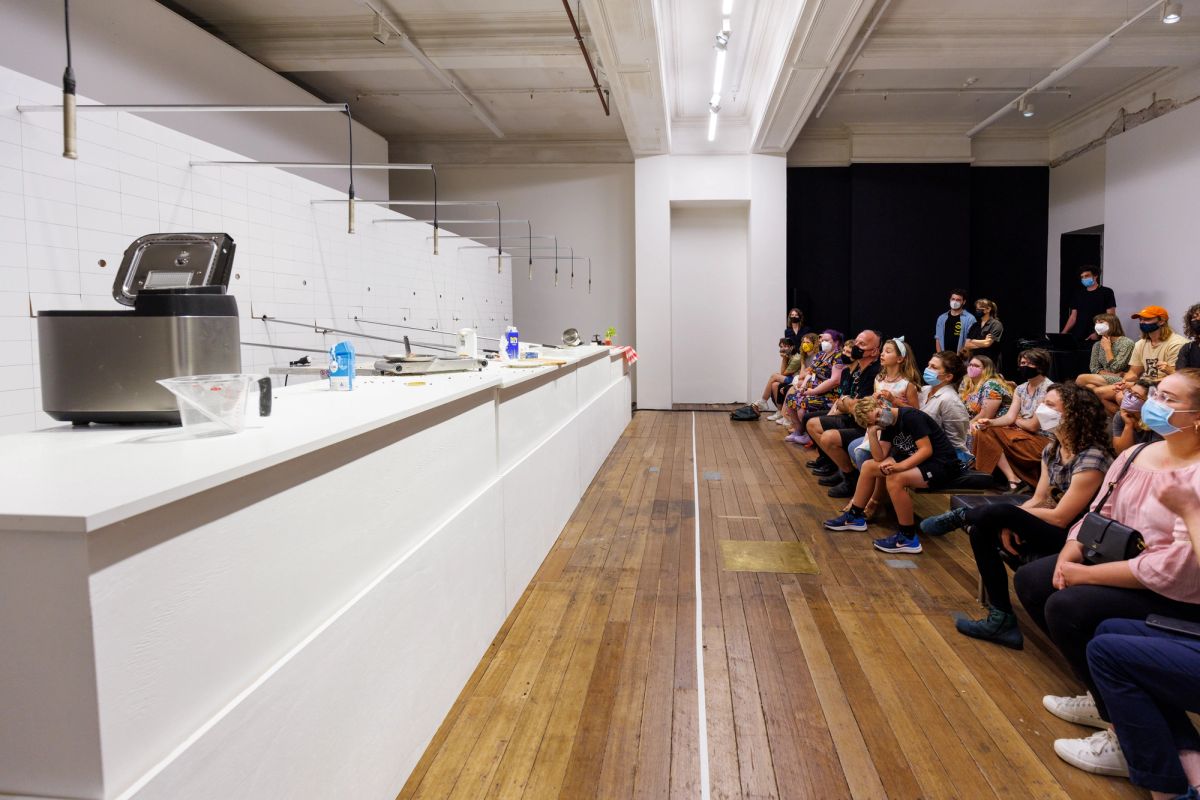 A group of people sit ready to watch All Day Breakfast, they're on seats, and opposite a white wall that has poles sticking out from it and breakfast paraphernalia on a table