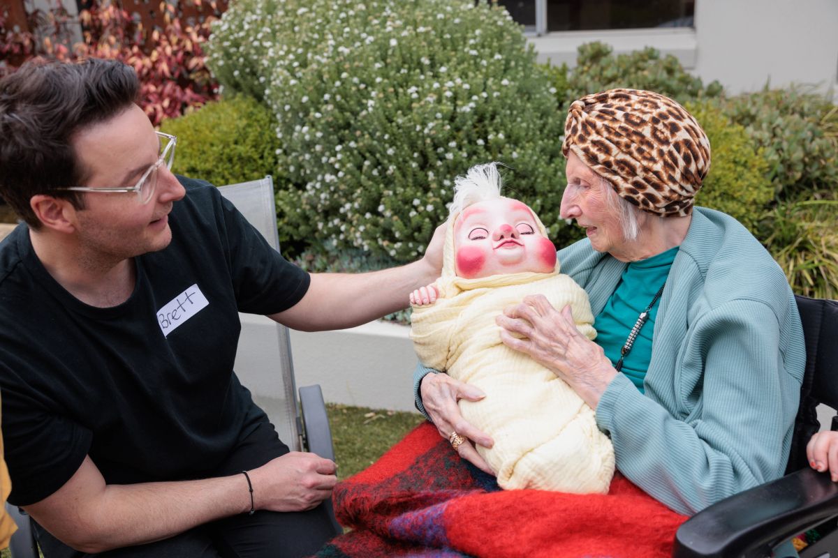 Two people sit looking at one another, one on the left wears a black t-shirt and has a name tag, saying 'Brett' the other person is elderly and in a chair, they hold a baby puppet wrapped in a yellow swaddle