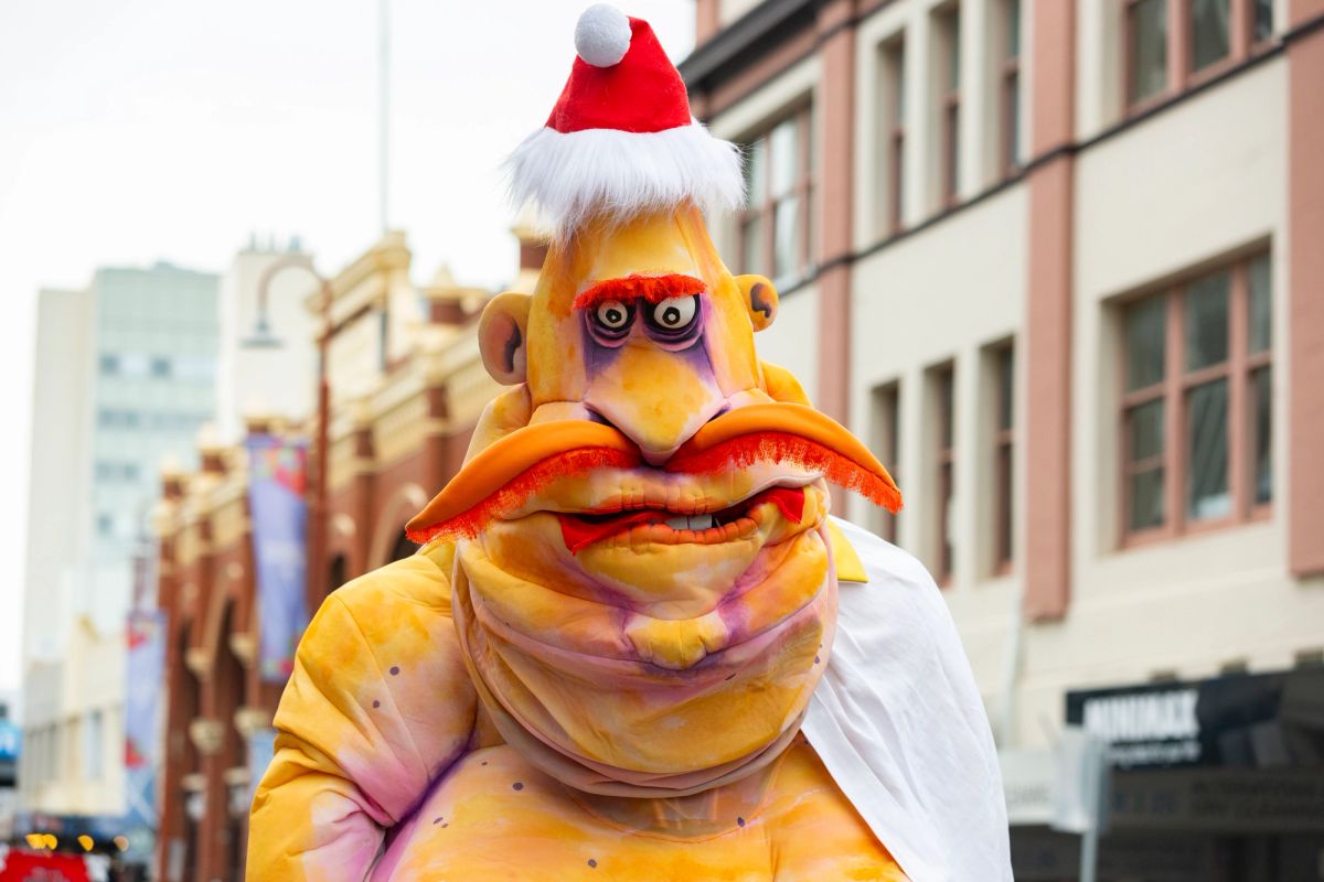 This is a close up image of Pa Ubu, a giant roving puppet, we see the puppet is wearing a Christmas hat, has over exaggerated features and a big orange moustache, he is walking in the City of Hobart Christmas Pageant, 2024, so in the background we see the tops of buildings.