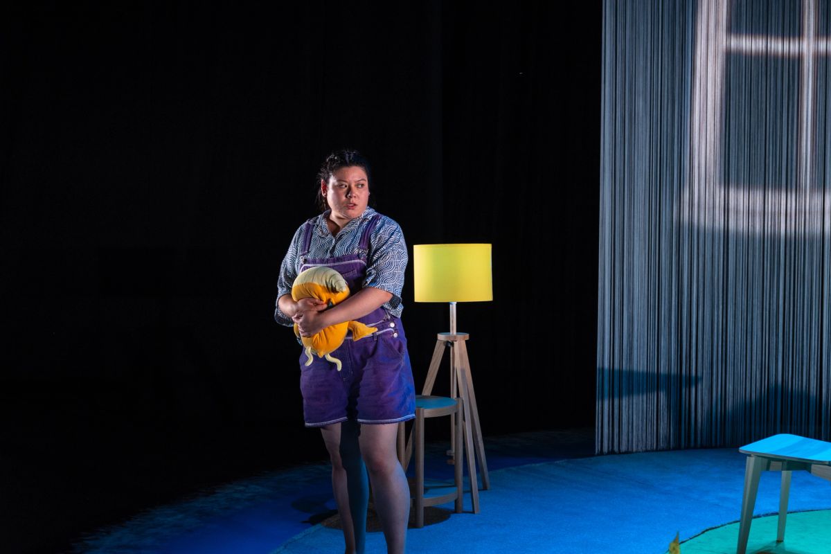 A performer dressed in purple overalls stands under stage lighting, holding a small yellow plush toy. She represents the daughter, Chi, in The Story of Chi, performed at Adelaide Festival Centre as part of OzAsia Festival 2024.