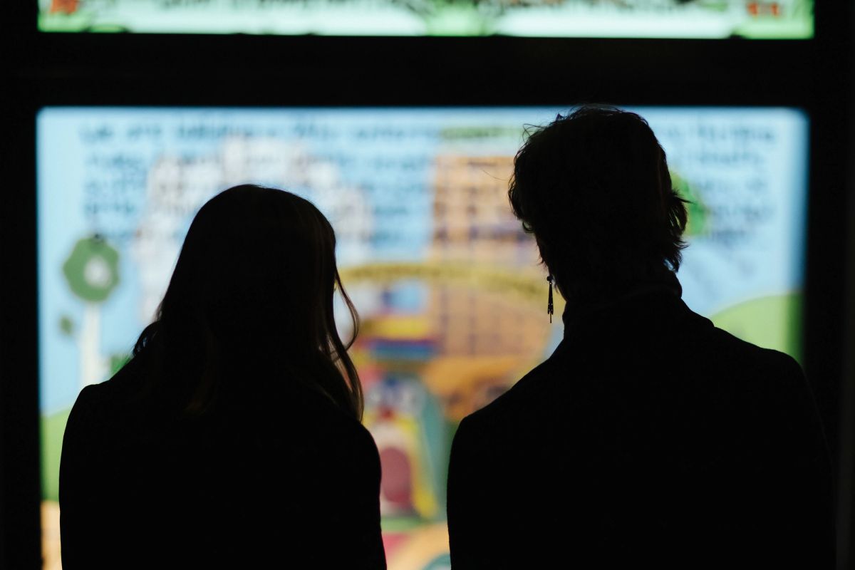 In a shop window we see a comic split into four parts, we look at the seemingly blurry comic over the shoulders of two adults reading it, they are silhouettes. The comic is colourful and a parrot features.
