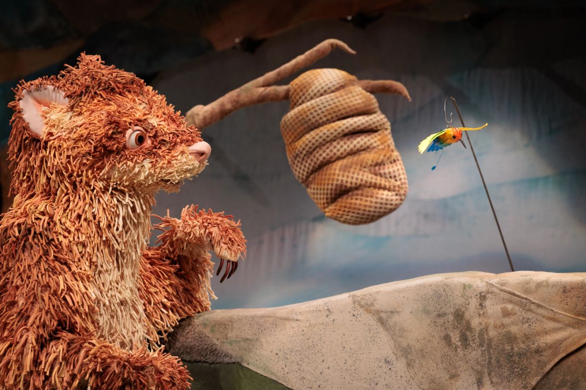 Feathers, a feathery-tailed ferret puppet, looks toward Rita the Bee Eater, a brightly coloured small puppet, made from lighting gels, milk cartons, and a knitting needle. With a beehive hanging behind them in Feathers at Theatre Royal Hobart.
